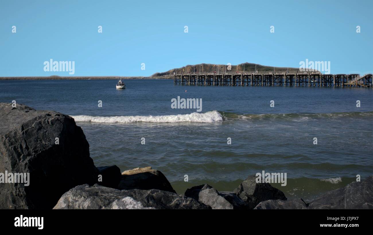 Jetty or Pier over sea with rocks, waves, yacht, boat, hill far in the distance. Coffs Harbour Jetty in Australia. Stock Photo