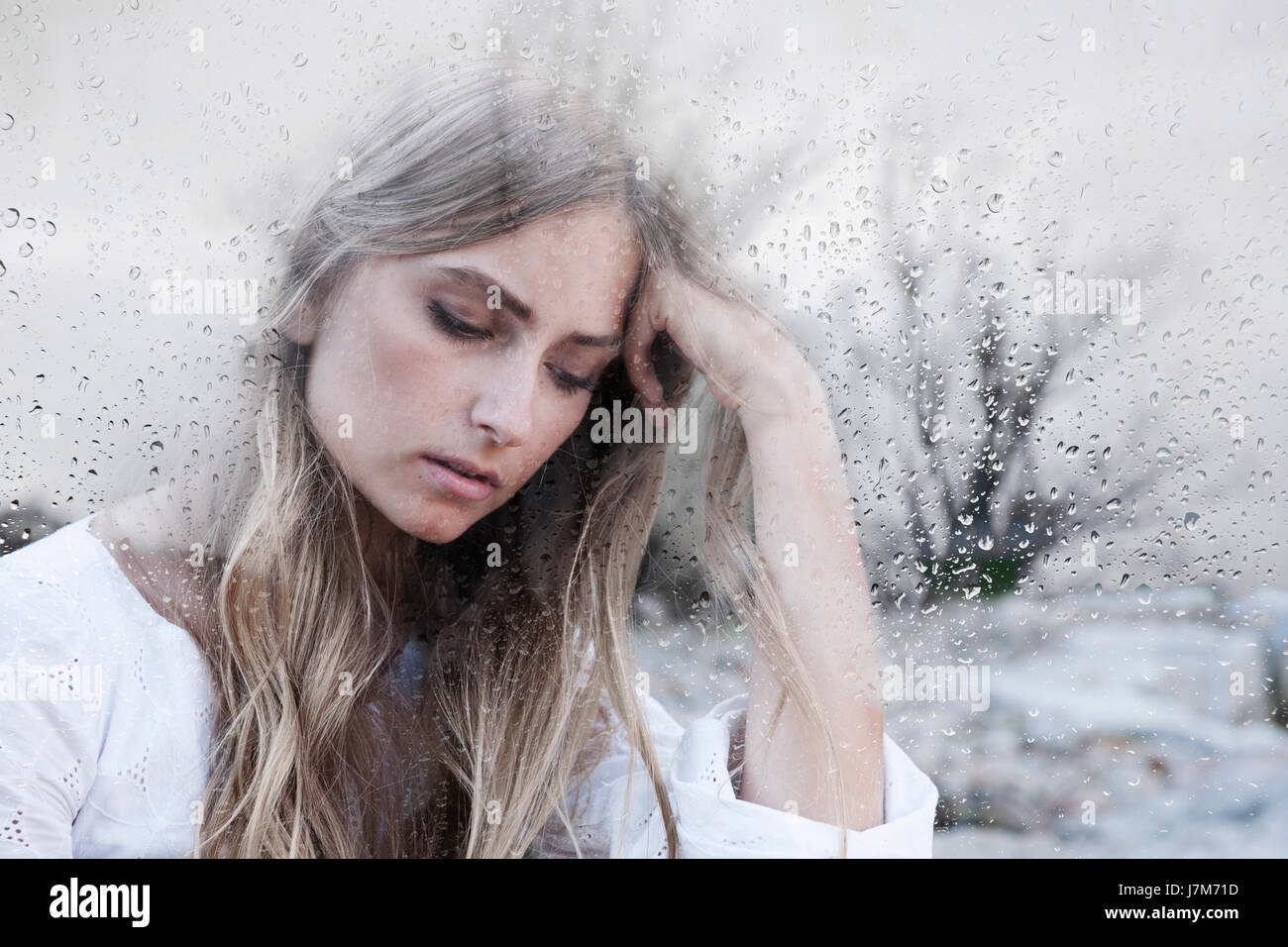 Young woman behind window glass on gray rainy day feeling sad Stock Photo