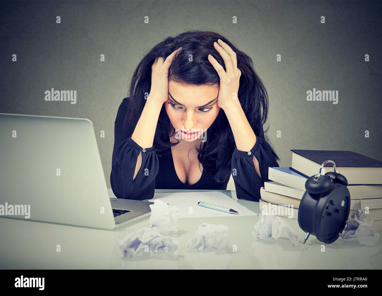 Too much work stressed woman sitting at her desk with books in front of laptop isolated gray office wall background. Stock Photo