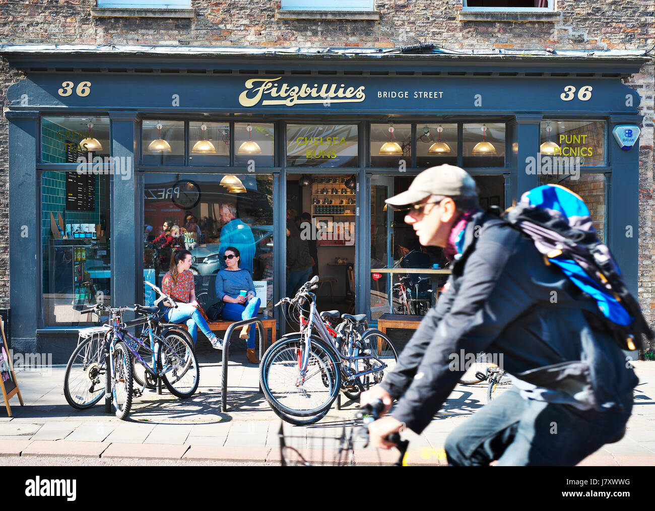 Sunday lifestyle in Cambridge, UK. Cyclist rides past a Cambridge institution of Fitzbilllies cafe in city while people sit drinking coffee Stock Photo