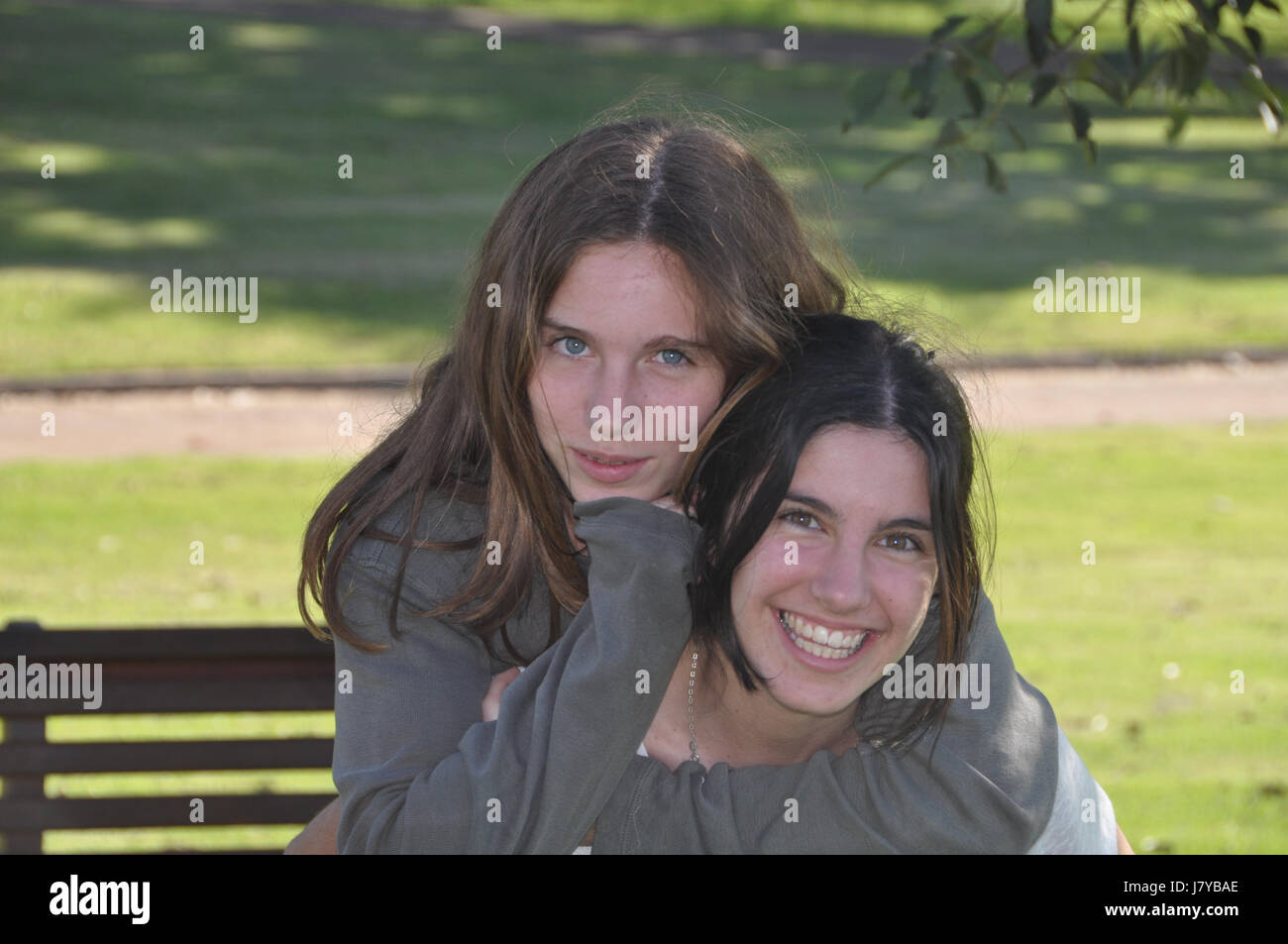 2 Teenage girls warm piggy back hug in shade on park bench with green background on sunny day Stock Photo