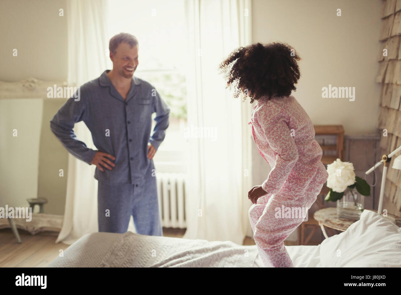 Multi-ethnic father in pajamas watching daughter jumping on bed Stock Photo