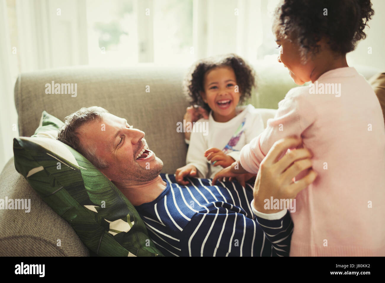 Playful multi-ethnic father and daughters tickling and laughing on sofa Stock Photo