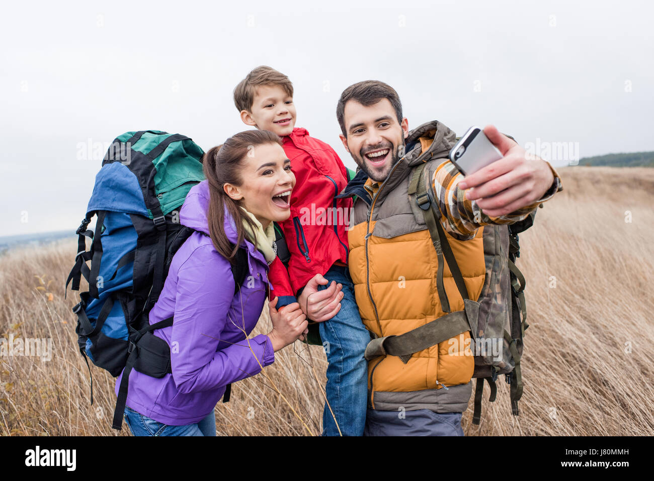 Happy family with backpacks standing in tall grass and taking selfie at autumn day Stock Photo