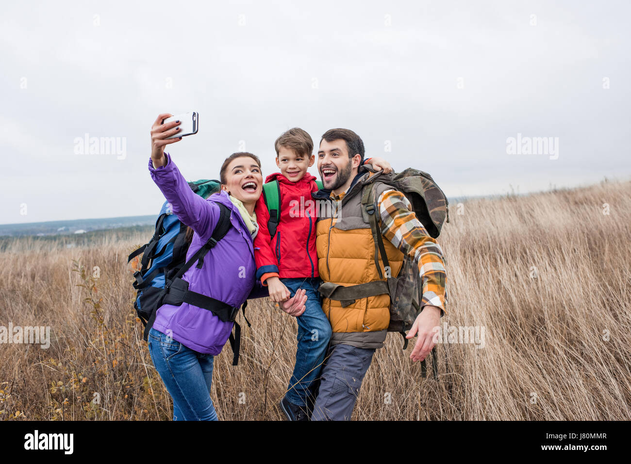 Happy family with backpacks standing in tall grass and taking selfie at autumn day Stock Photo