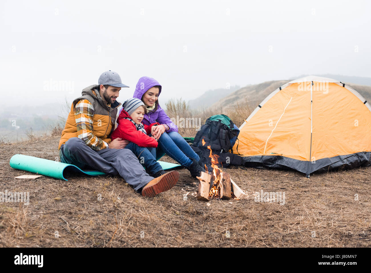 Happy family sitting near tent and looking at burning fire at cloudy autumn day Stock Photo