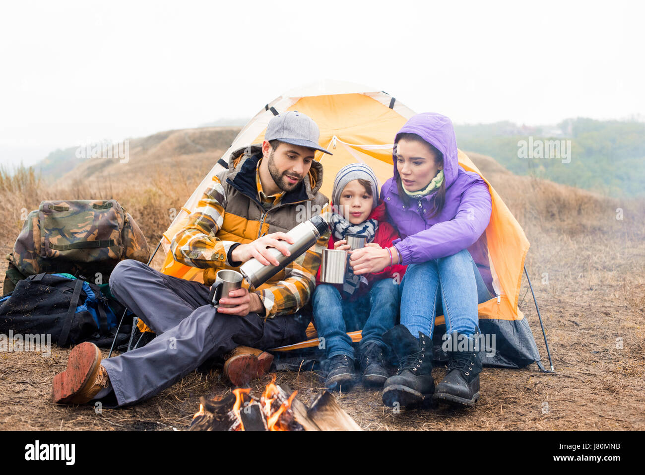 Family with one child sitting near tent and drinking hot tea from thermos while resting near burning fire in countryside Stock Photo