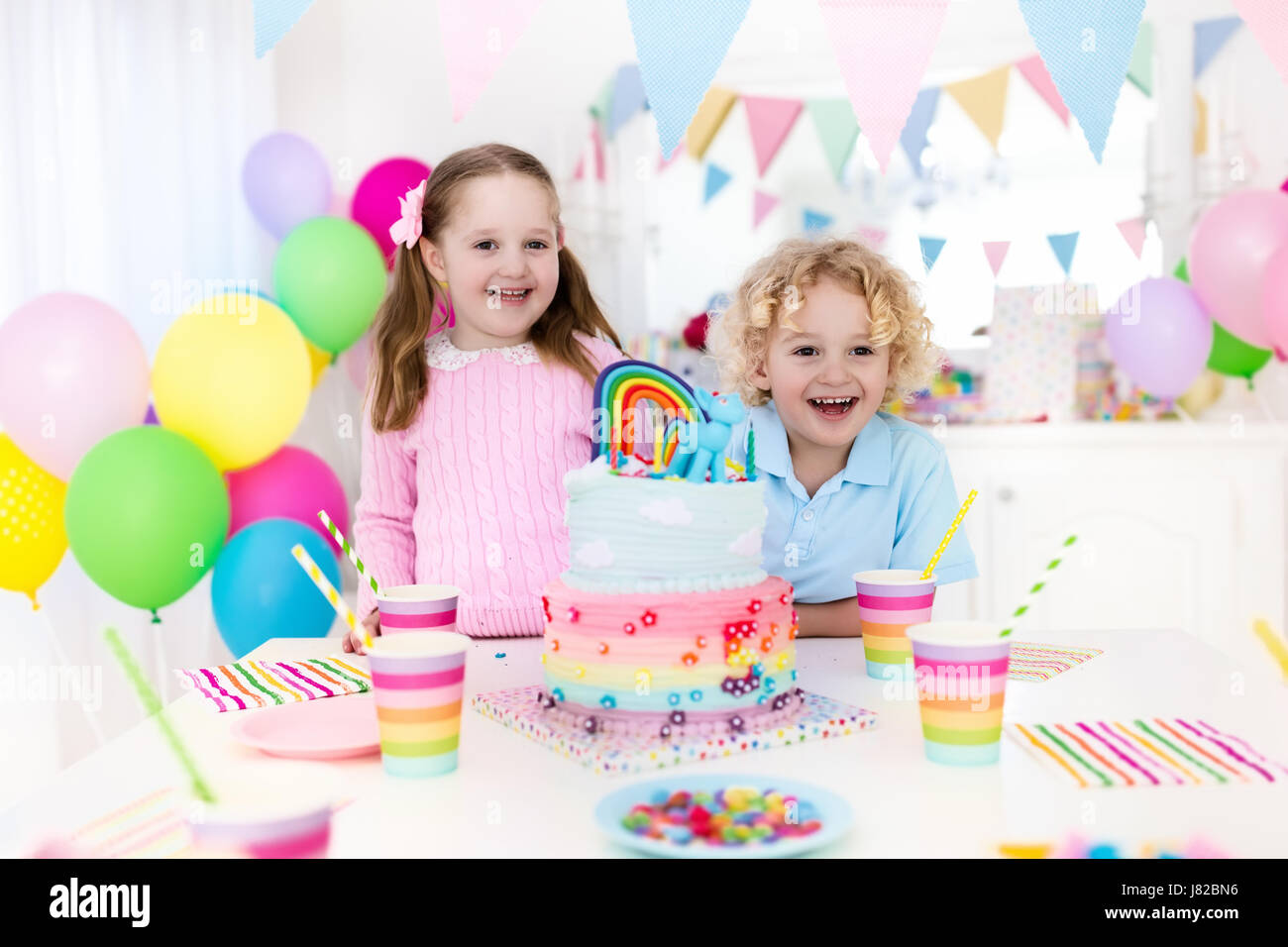 Kids birthday party with colorful pastel decoration and rainbow cake. Girl and boy with sweets, candy and fruit. Balloons and banner at festive decora Stock Photo