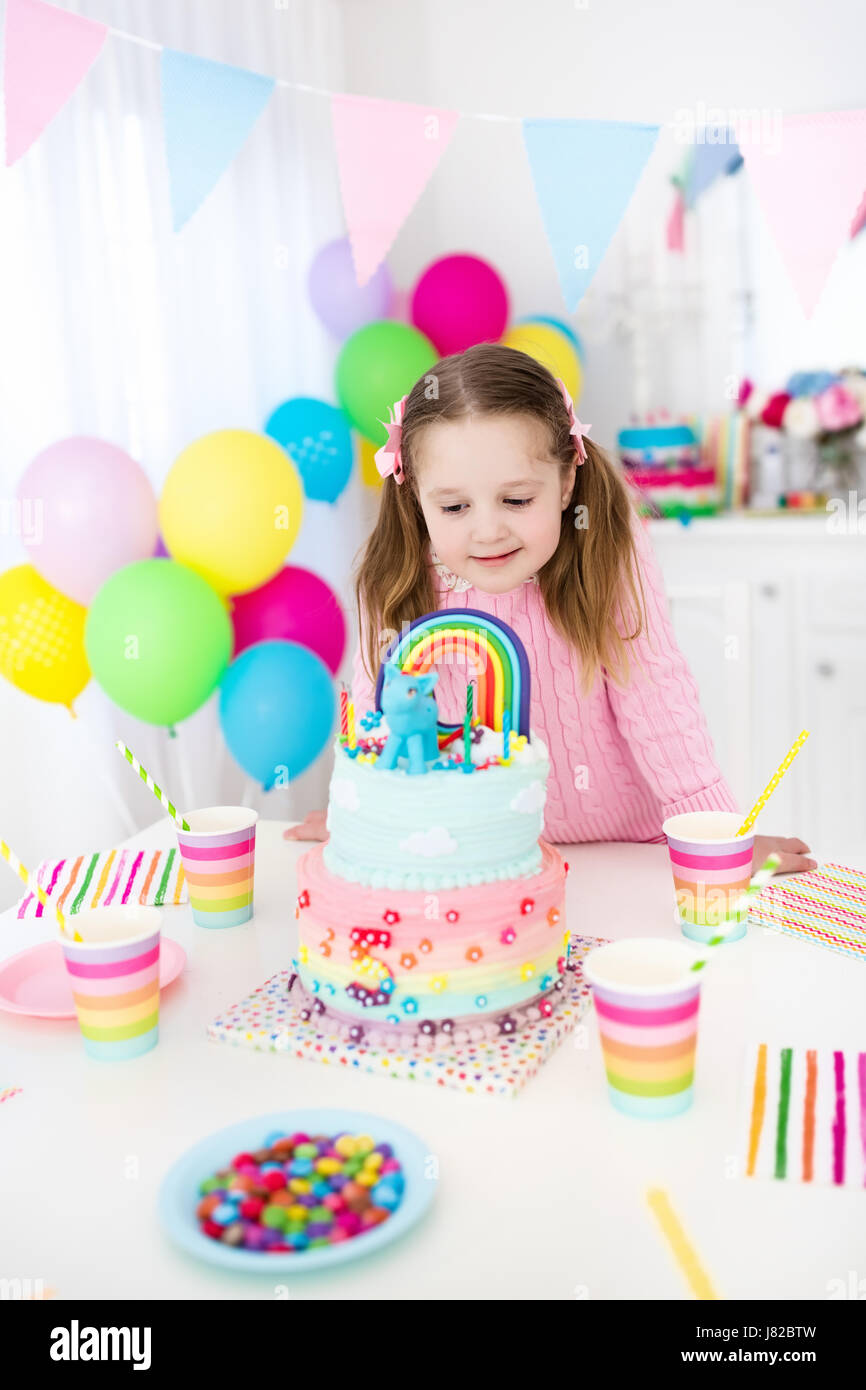 Kids birthday party with colorful pastel decoration and unicorn rainbow cake. Little girl with sweets, candy and fruit. Balloons and banner at festive Stock Photo