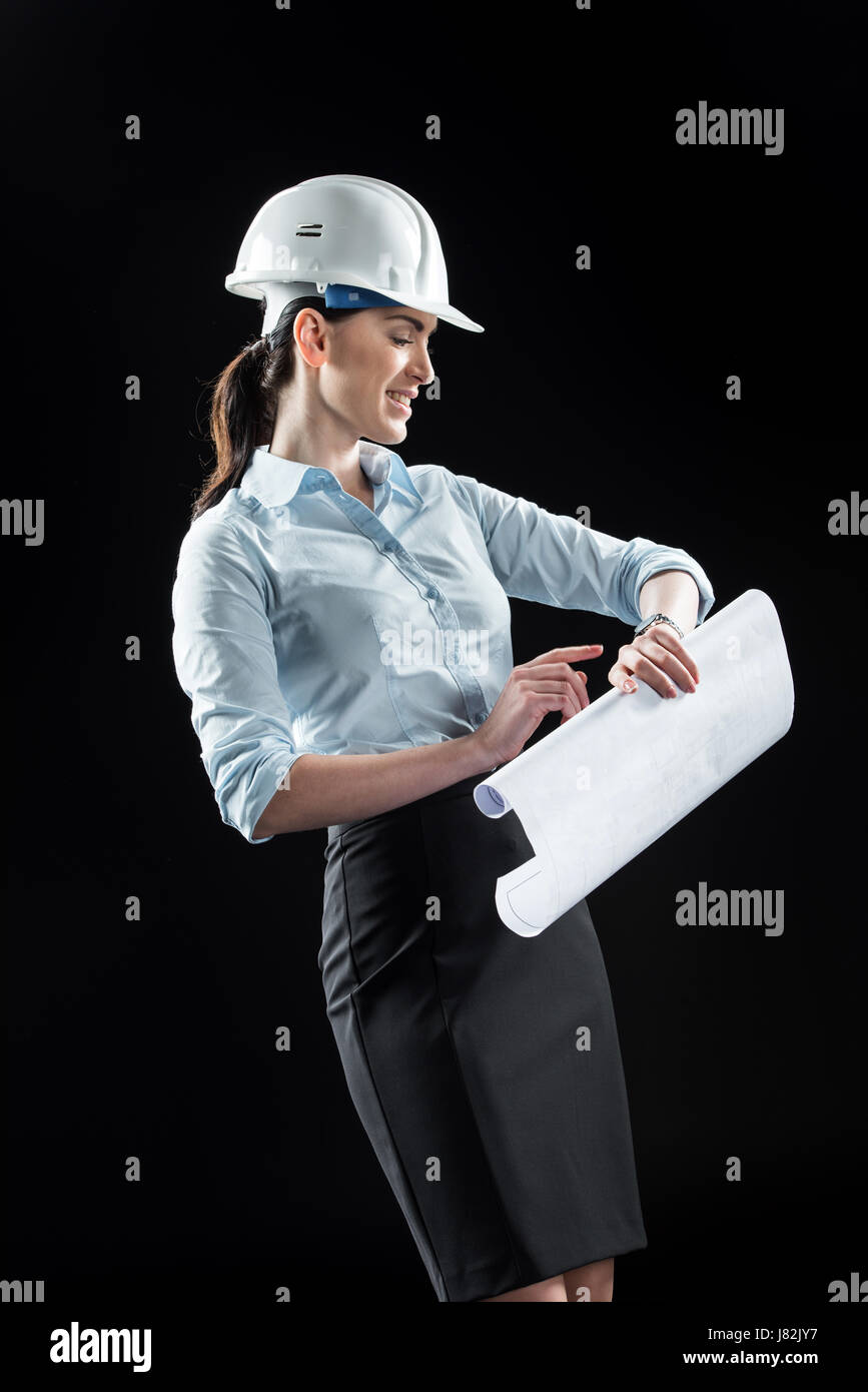 Smiling female architect in helmet holding blueprint and looking at ...