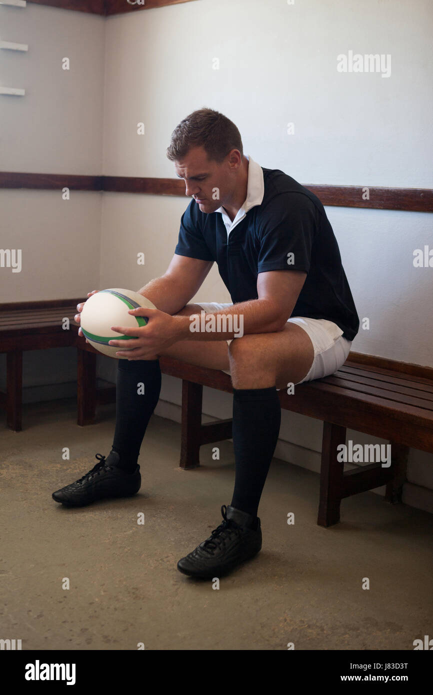 Thoughtful rugby player holding ball while sitting on bench in locker ...