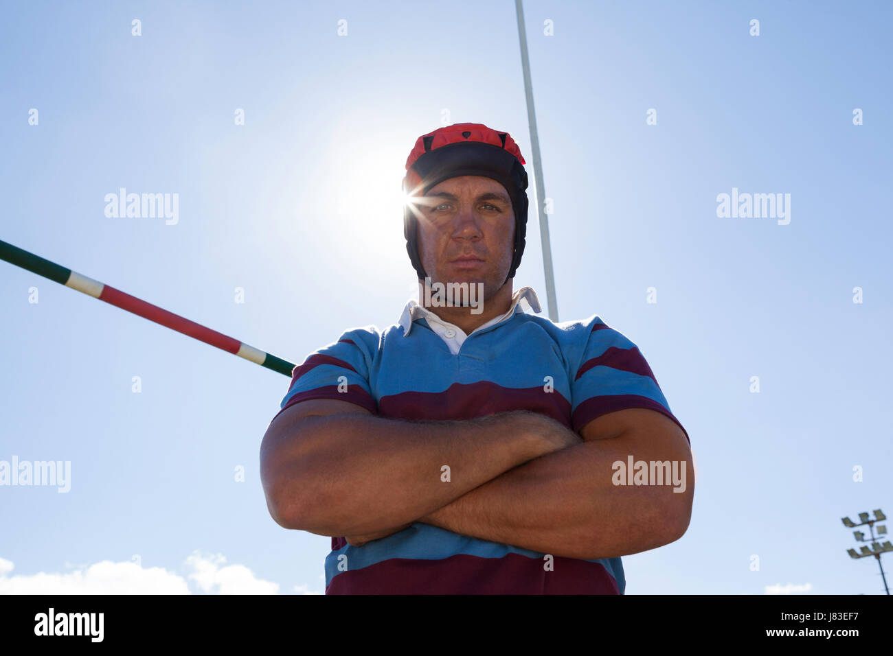 Low angle portrait of rugby player wearing helmet with arms crossed against blue sky on sunny