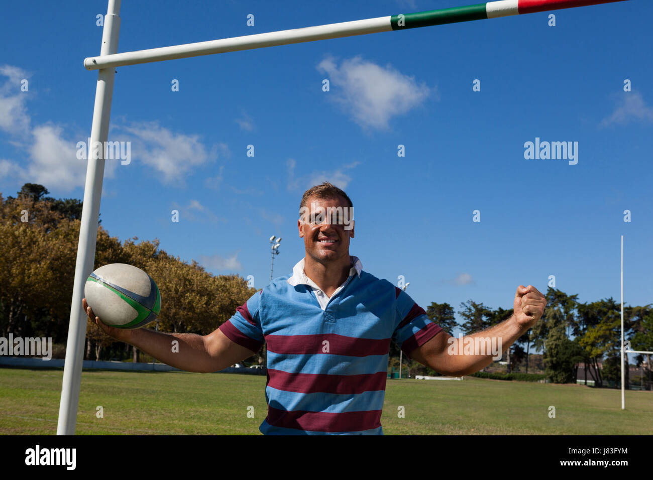 Portrait of happy rugby player holding ball by goal post against blue sky Stock Photo