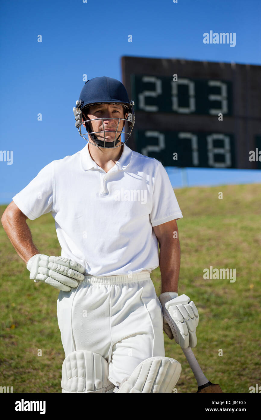 Confident cricket player with bat standing against scoreboard at field