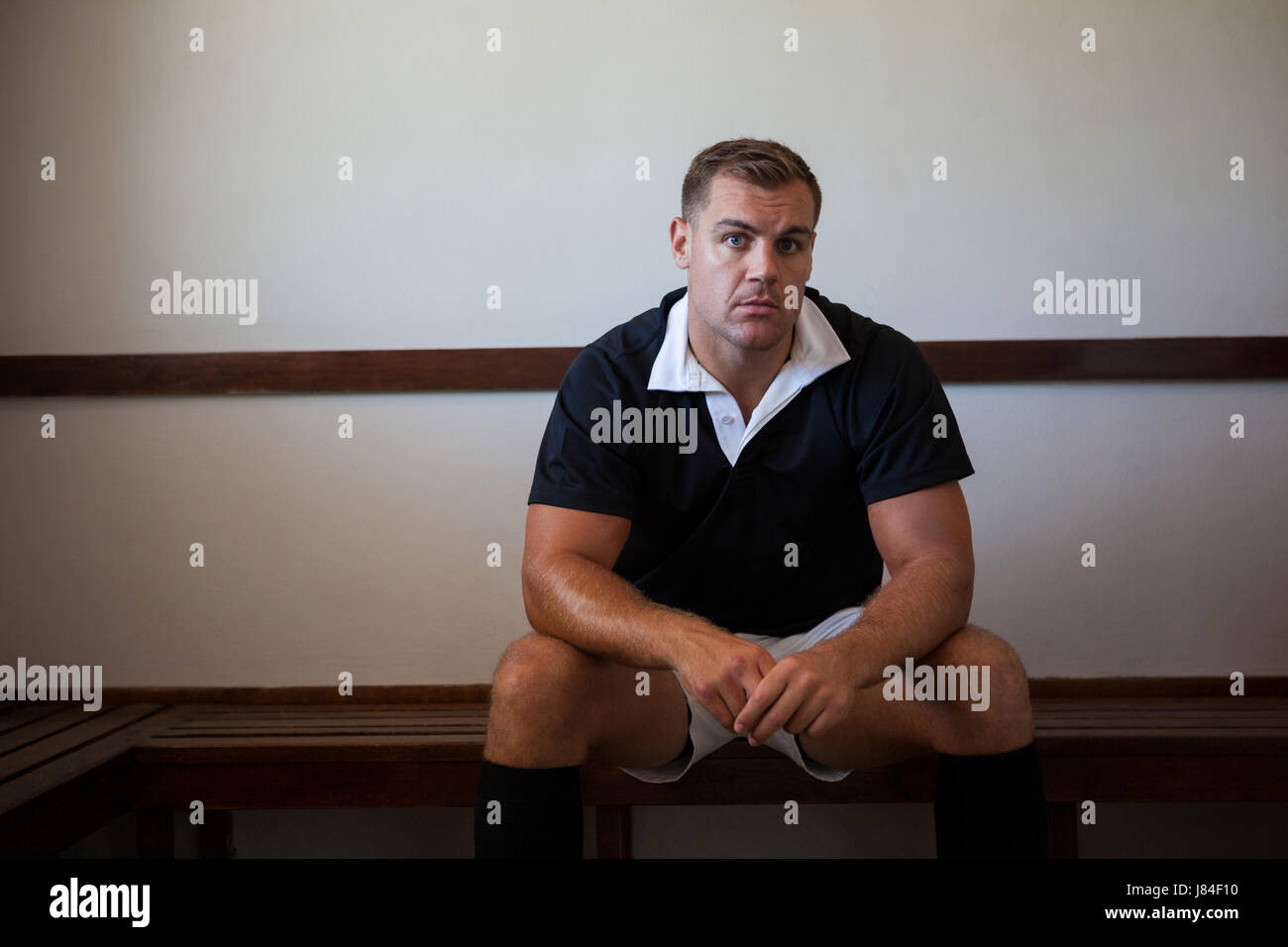 Portrait of serious rugby player sitting on wooden bench against wall ...