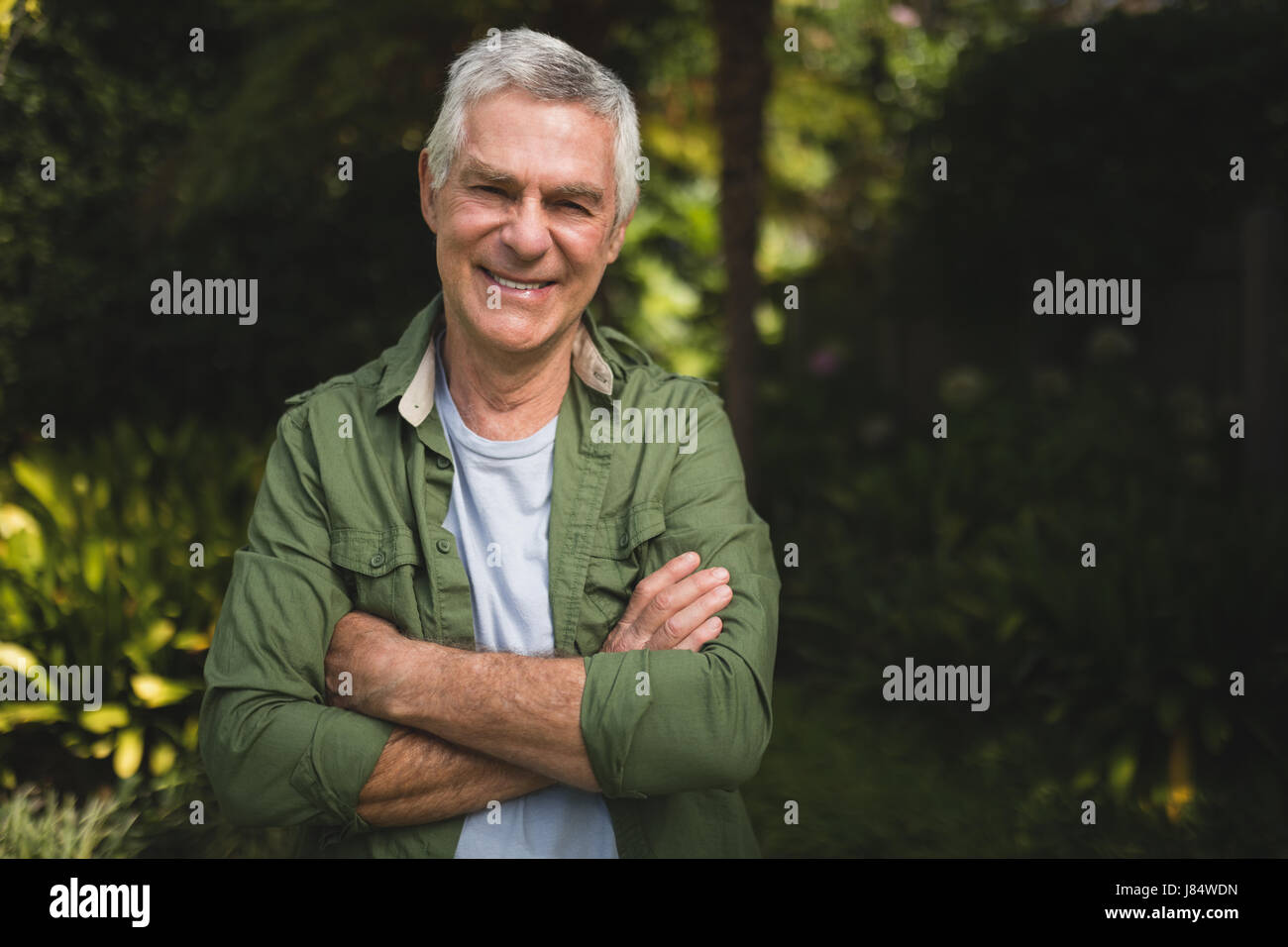 Portrait of smiling senior man standing with arms crossed in backyard ...