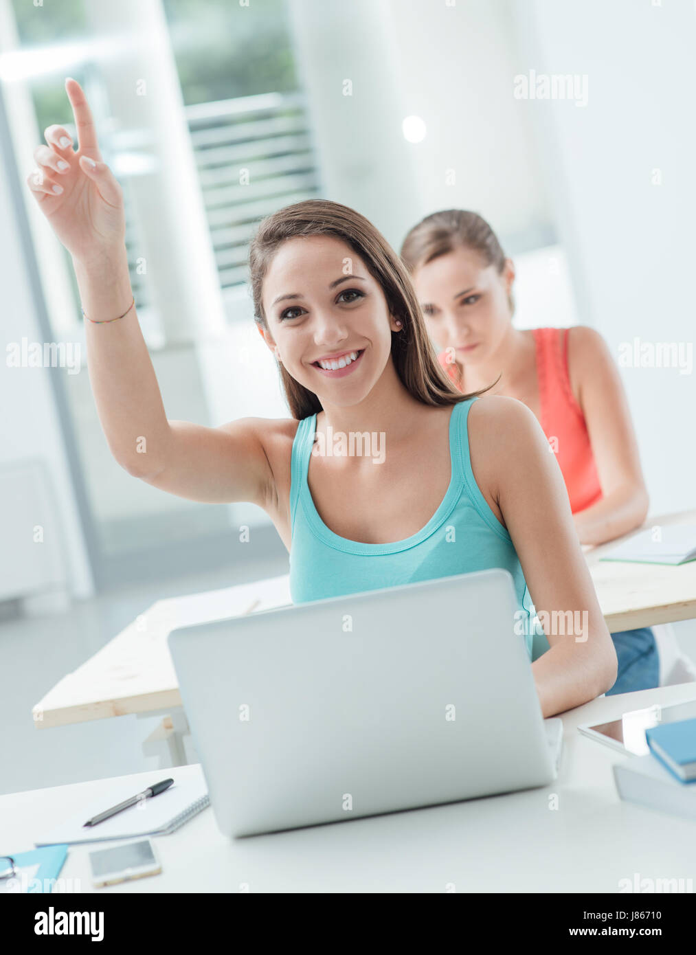 Smart smiling girl sitting at school desk and raising her hand to ask ...