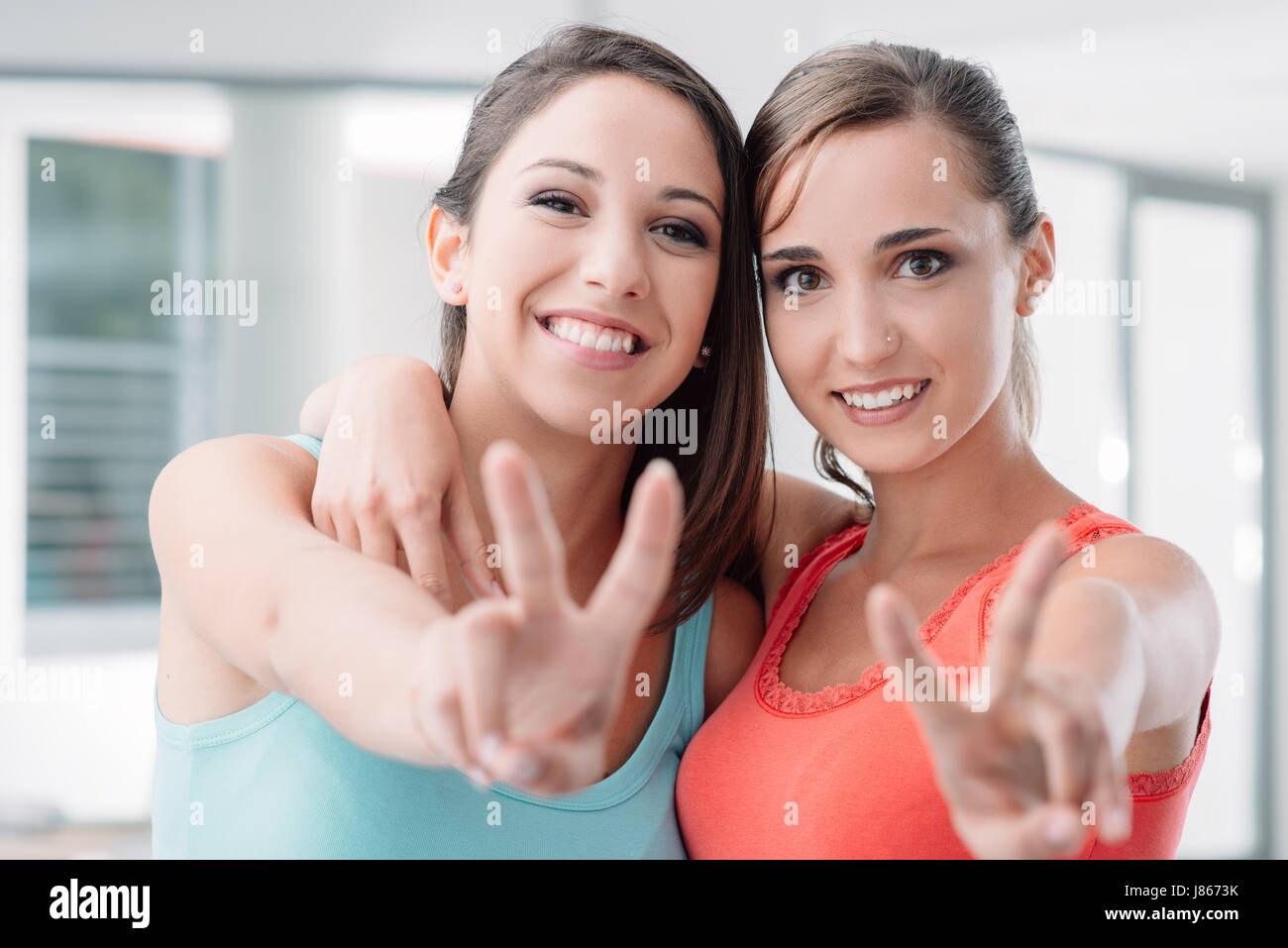 Cheerful teen girls posing, making a V sign and smiling at camera, one is hugging her friend Stock Photo