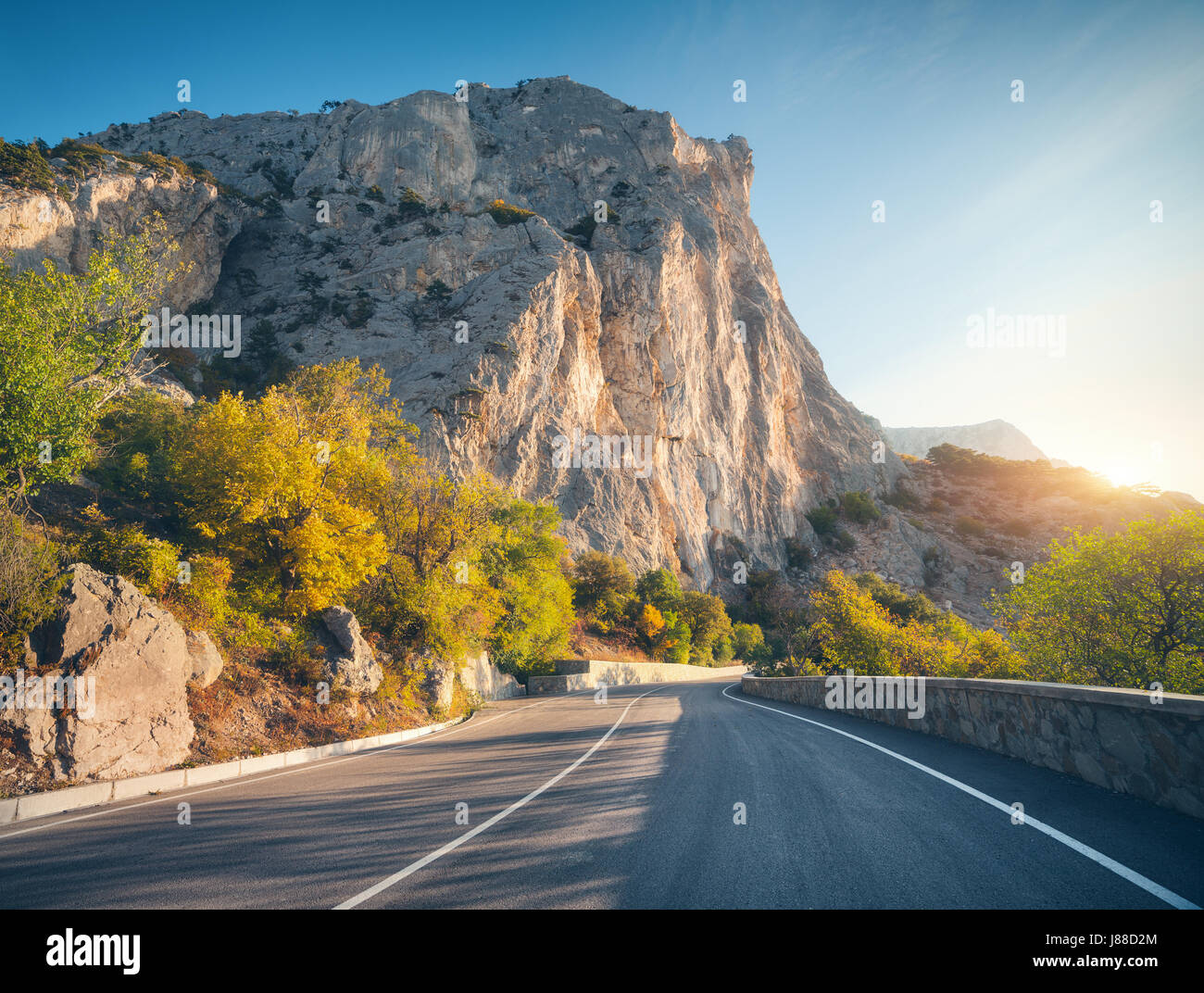 Asphalt road in summer at sunrise. Landscape with beautiful empty mountain road with a perfect asphalt, high rocks, trees and sunny blue sky. Travel b Stock Photo