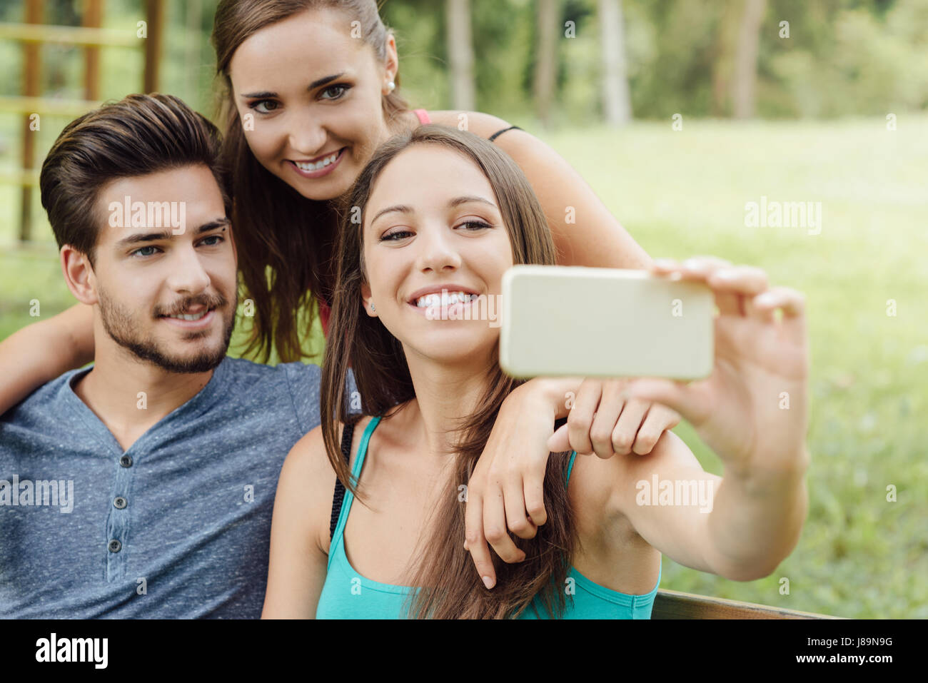 Cheerful smiling teens at the park sitting on a bench and taking selfies using a smart phone Stock Photo