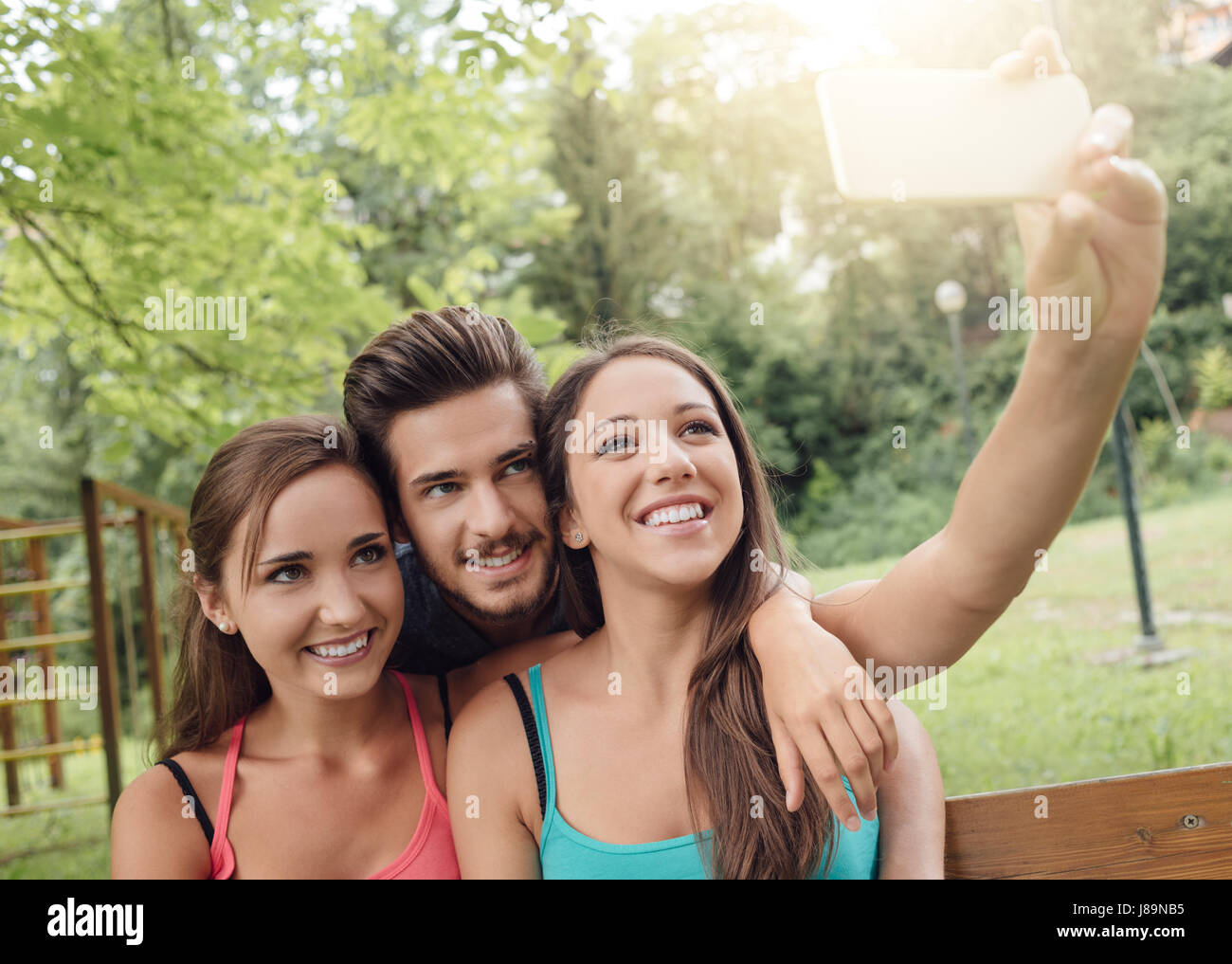 Cheerful smiling teens at the park sitting on a bench and taking selfies using a smart phone Stock Photo