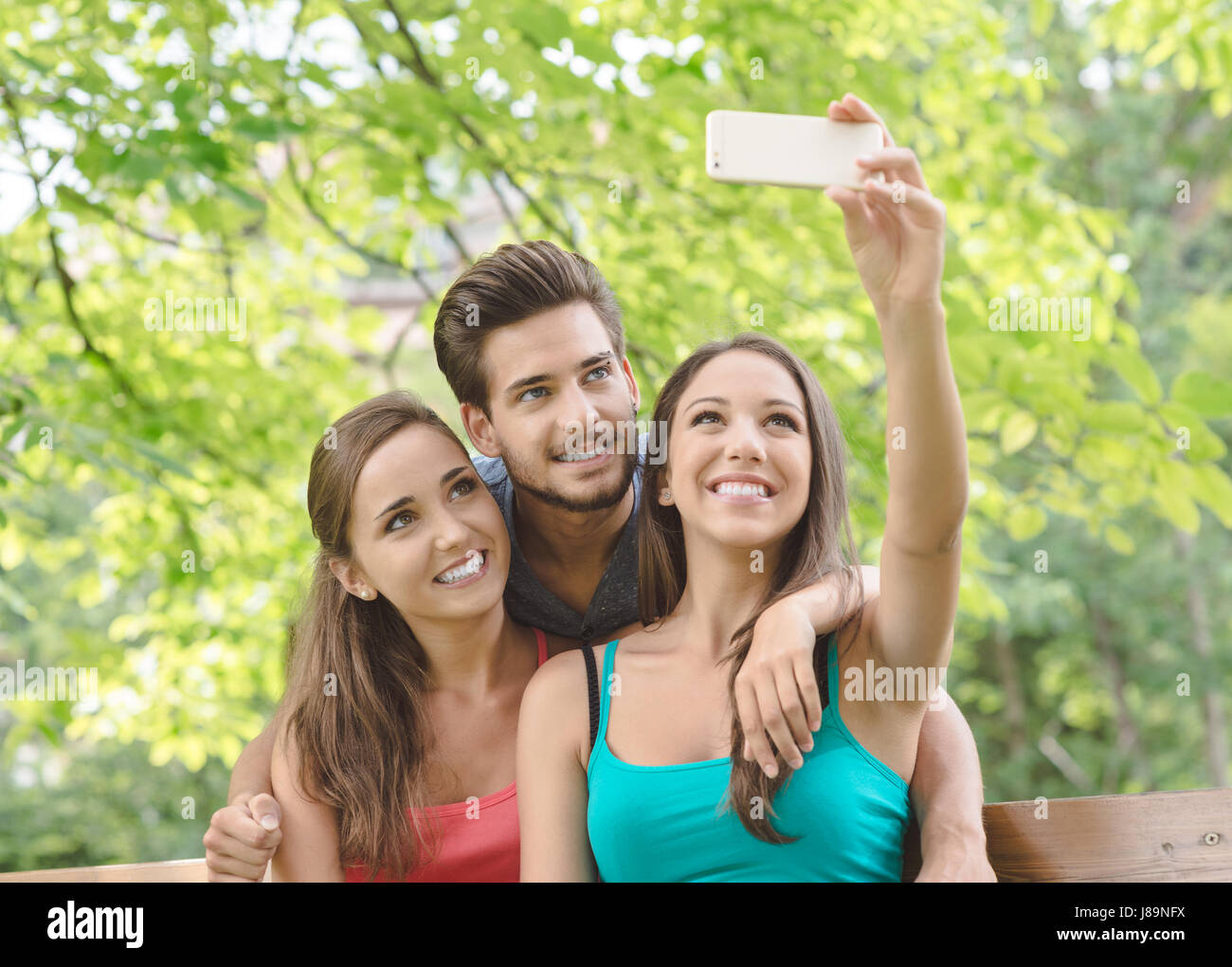Cheerful smiling teens at the park sitting on a bench and taking selfies using a smart phone Stock Photo