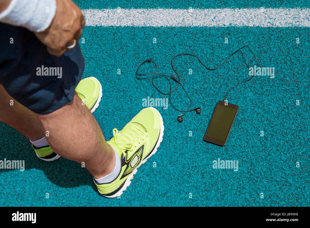 Male legs with running sneakers, smartphone with blank screen and in ear headphones on blue running track. Top down point of view. Sport, technology,  Stock Photo