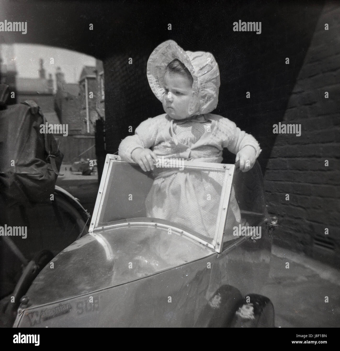 1950s, a pensive looking female toddler in her Sunday best bonnet standing on the seat of a British made 'Watsonian Sub', a child or juvenile sidecar that fitted onto the side of a pedal bicycle. Taking young children in small trailers such as these, was popular in this ear before the motorcar became affordable for the masses. Watsonia sidecars were made in Albion Rd, Greet, Birmingham, England, UK. Although made primarily for bicycle use, they were also fitted to small motorccycles, such as mopeds. Stock Photo