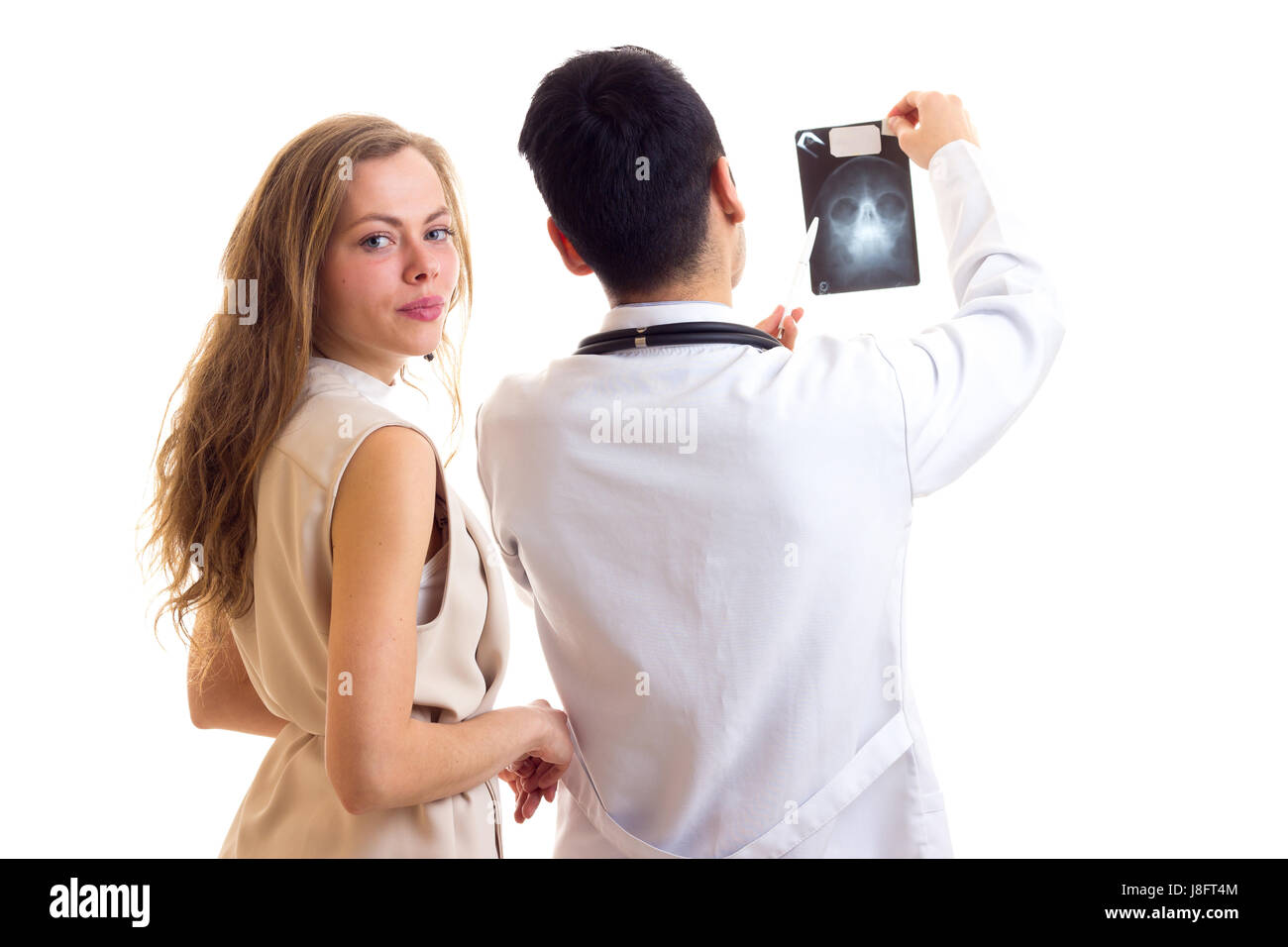 Young doctor with black hair in white gown with stethoscope showing X-ray to young charming woman with long brown hair in beige dress on white backgro Stock Photo