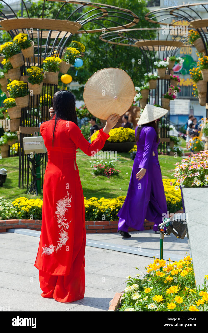 Ho Chi Minh City, Vietnam - February 07, 2016: Two young Vietnamese women in traditional Ao Dai dress are posing for pictures in front flower beds at  Stock Photo