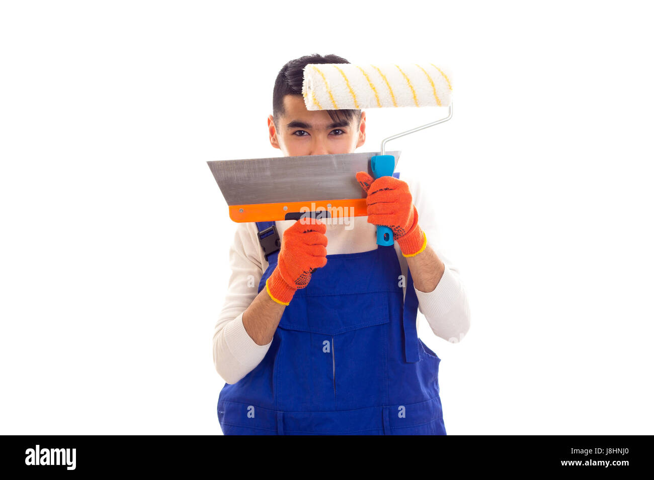 Young man with dark hair in white shirt and blue overall with orange gloves holding spatula and roll on white background in studio Stock Photo