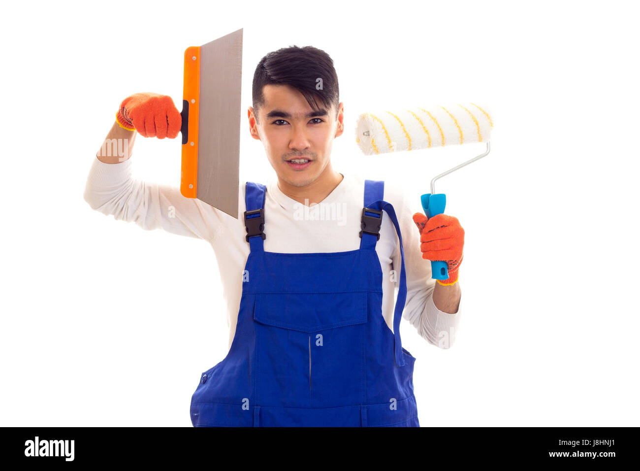 Young arrogant man with dark hair in white shirt and blue overall with orange gloves holding spatula and roll on white background in studio Stock Photo