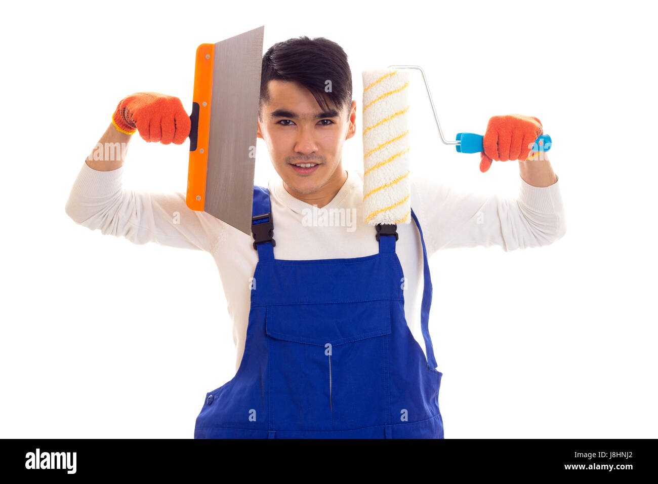 Young smartlooking man with dark hair in white shirt and blue overall with orange gloves holding spatula and roll on white background in studio Stock Photo