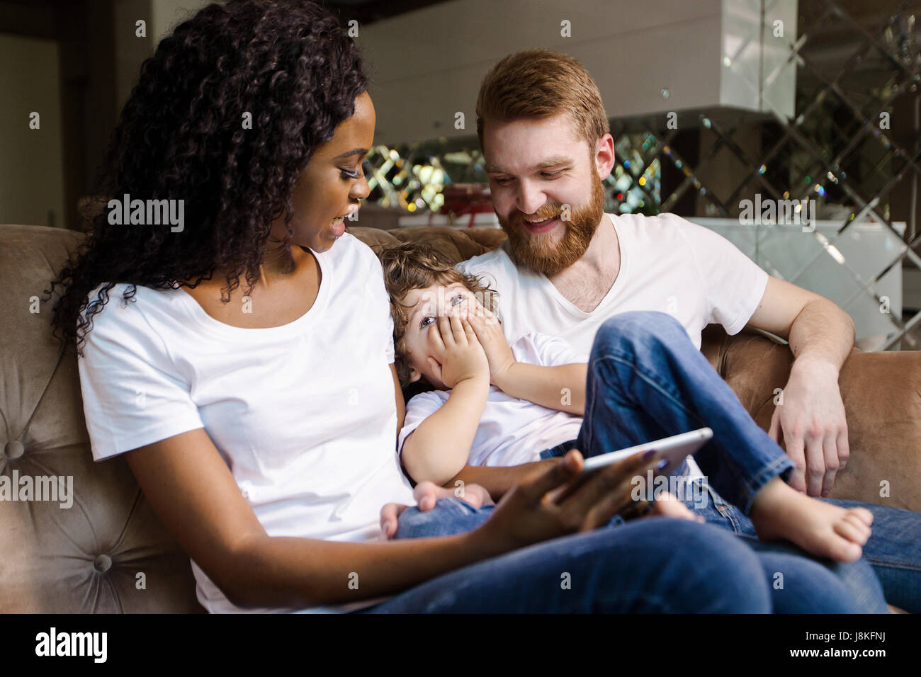 Happy couple of black woman and white man sitting on the sofa with their  son. Woman holding tablet Stock Photo - Alamy