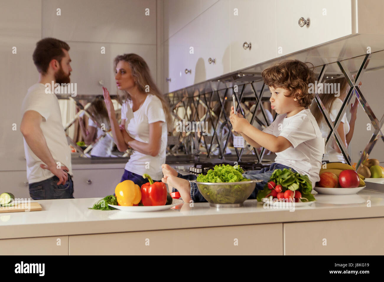 Family conflict photo. Little boy sitting on the kitchen surface while his parents fighting on the background. Stock Photo