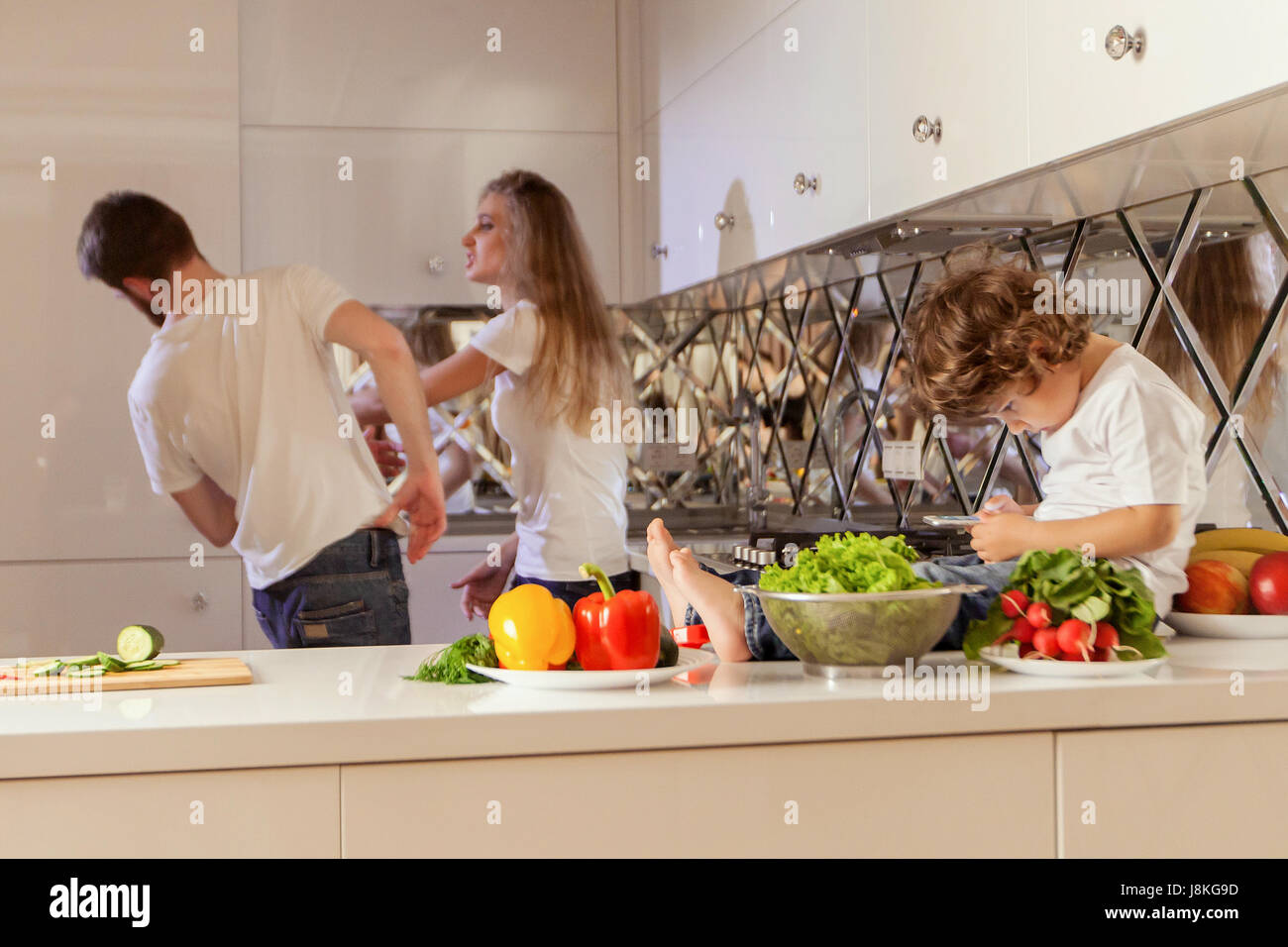 Family conflict. Small boy sitting on the kitchen surface with the phone. Parents fighting on the background. Stock Photo