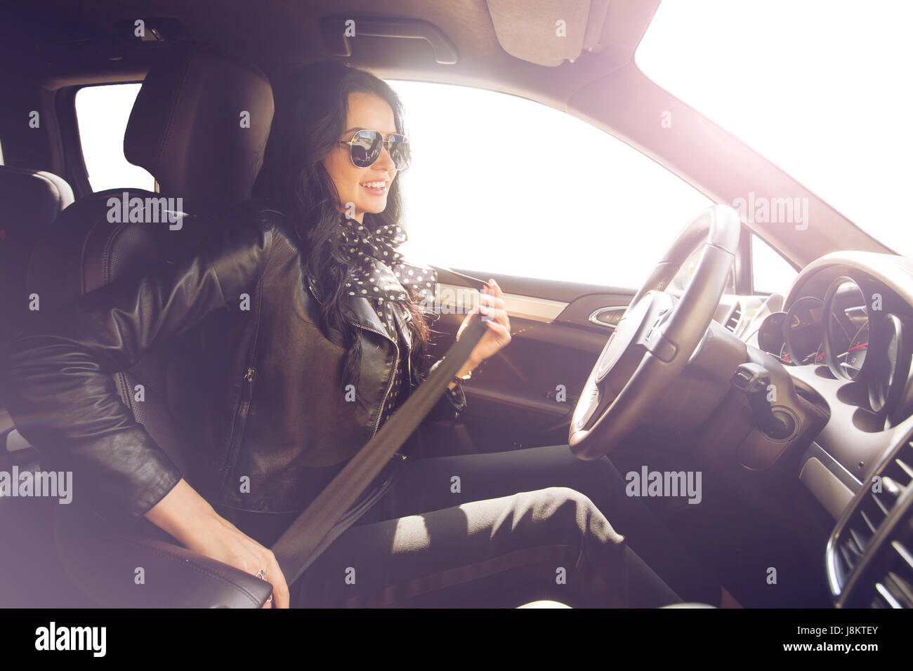 Brunette in sunglasses woman fastens with a belt in an expensive car. Safety first. Beautiful caucasian lady fastening car seat belt. Stock Photo