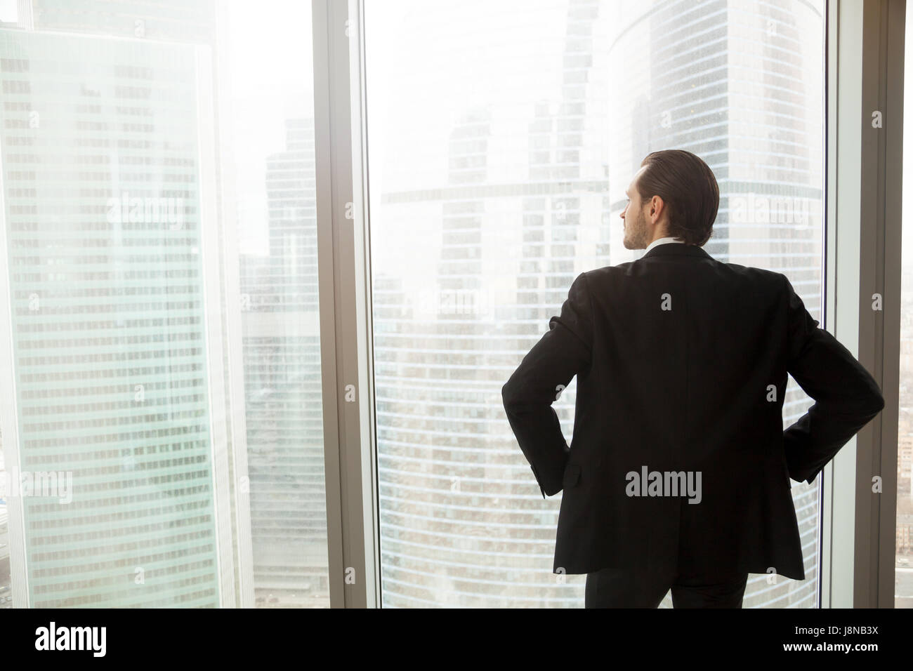 Back view of man in suit standing with hands on hips looking on cityscape outside the window. Successful businessman thinking about future plans, guy  Stock Photo