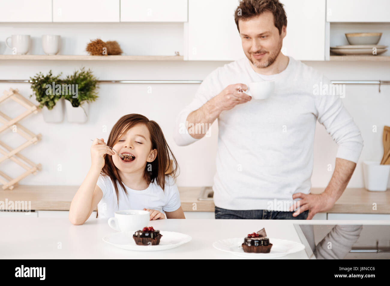 Pure delight. Blissful cute little girl relishing chocolate cake with her eyes closed and a happy smile while her father standing behind her and looki Stock Photo