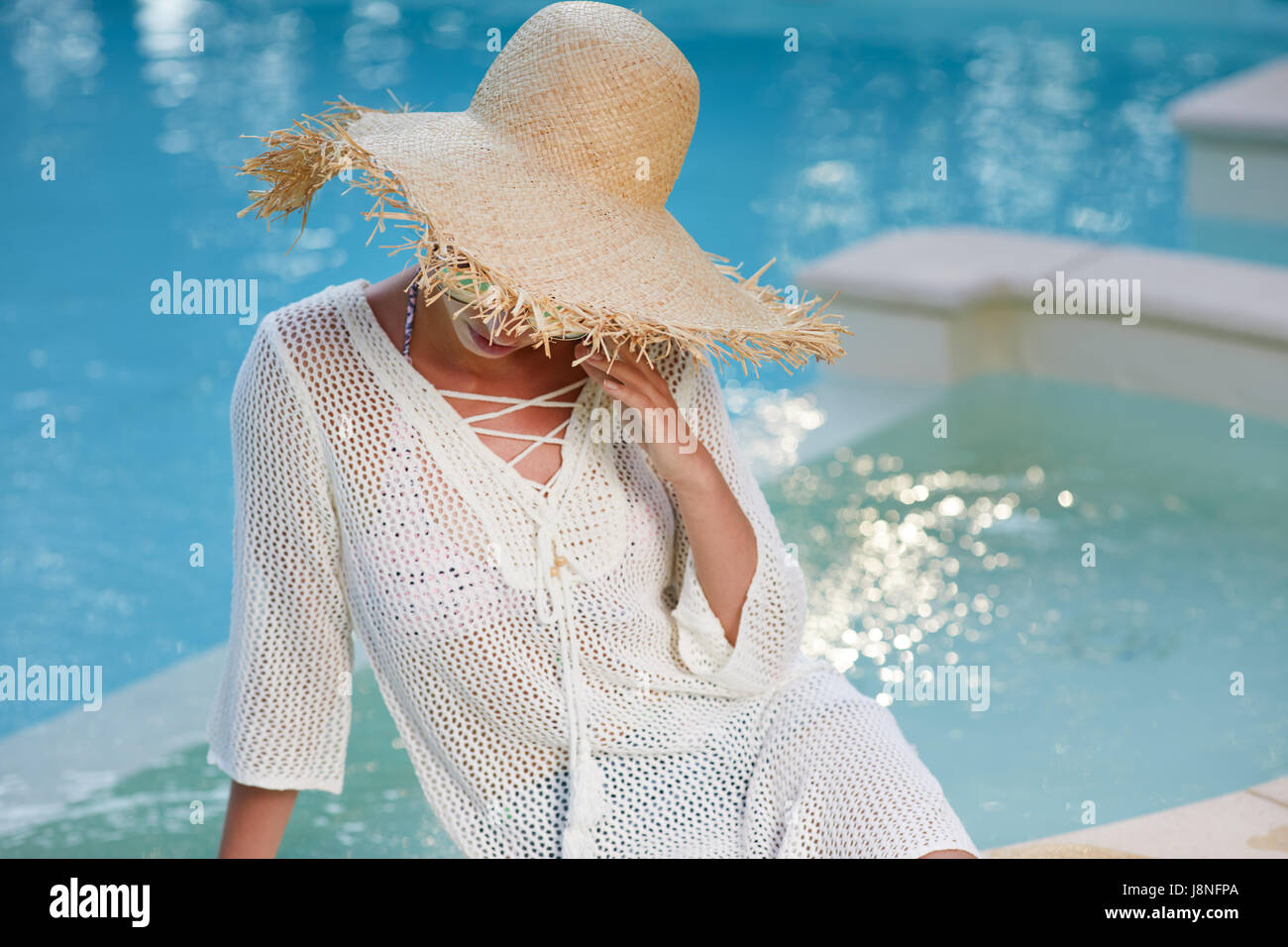 Beautiful girl in a  dress and sunhat sits next a resort pool Stock Photo