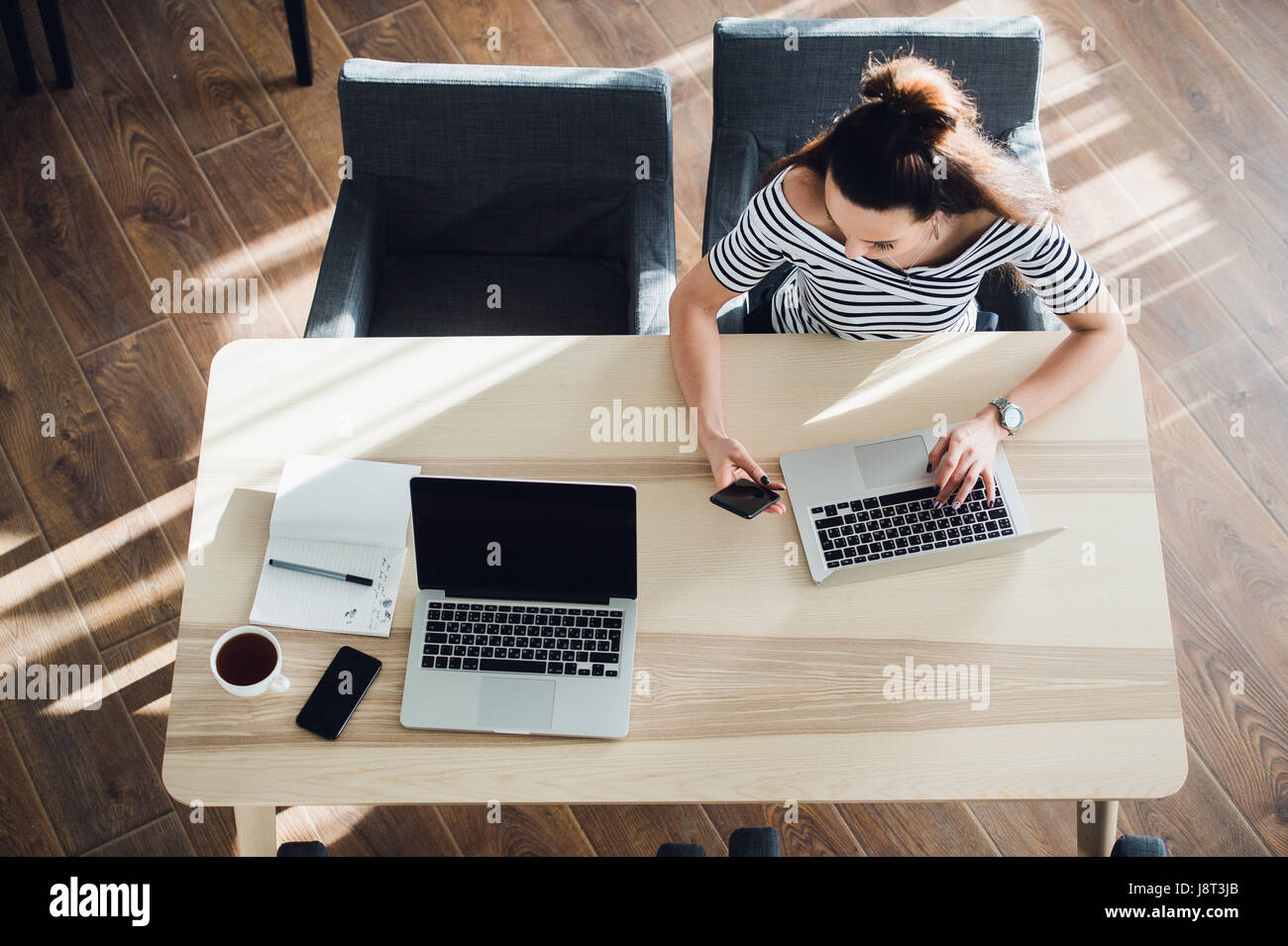 Busy young woman texting with mobile phone and writing notes while sitting at her desk. Pretty caucasian female working in home office or a cafe with her laptop. Stock Photo