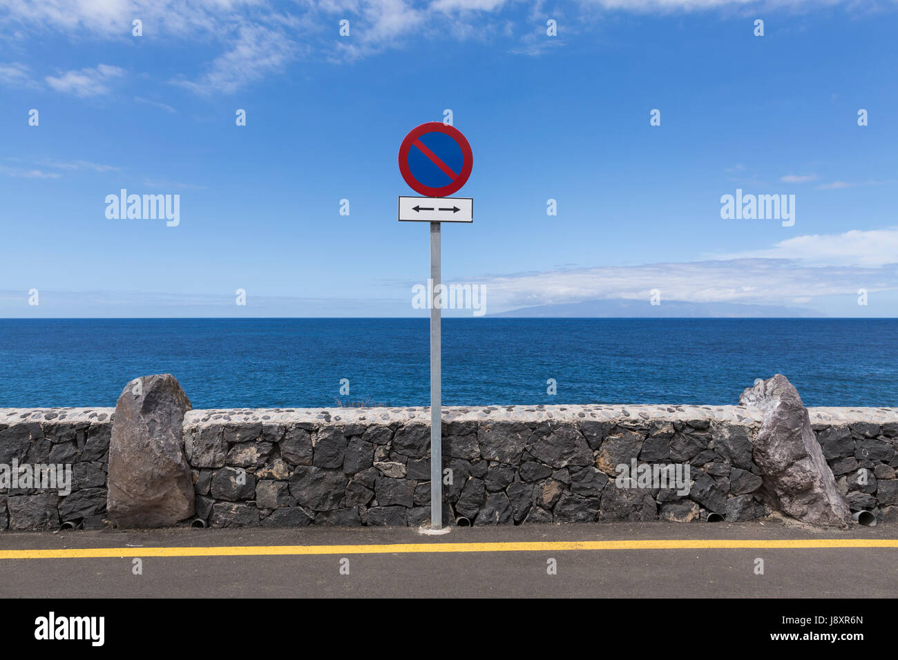 Yellow line on road and No parking sign next to natural stone wall at the coast with the atlantic ocean and the island of La Gomera in the background, Stock Photo