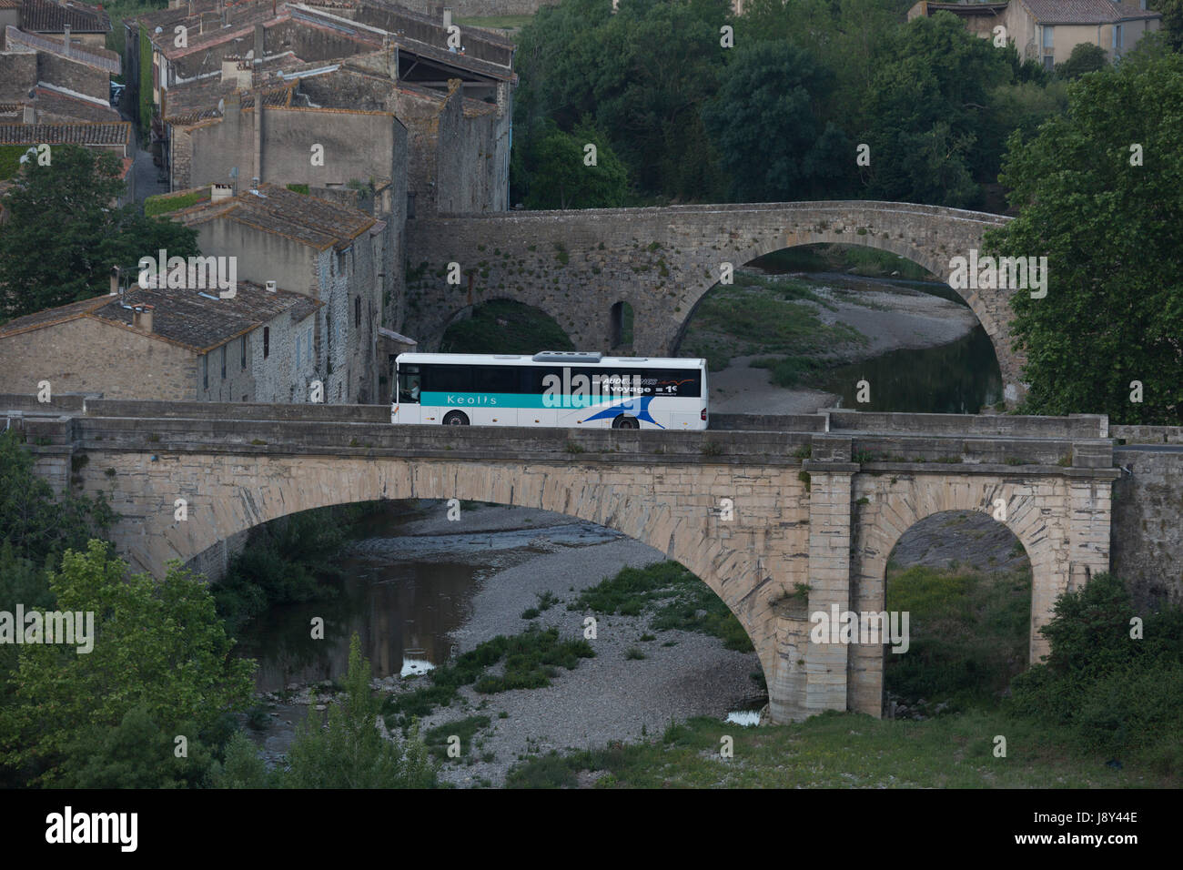 The early school bus crosses one of the bridges at the pretty French medieval walled village of Lagrasse on the River Orbieu, on 23rd May, 2017, in Lagrasse, Languedoc-Rousillon, south of France. Lagrasse is listed as one of France's most beautiful villages and lies on the famous Route 20 wine route in the Basses-Corbieres region dating to the 13th century. Stock Photo