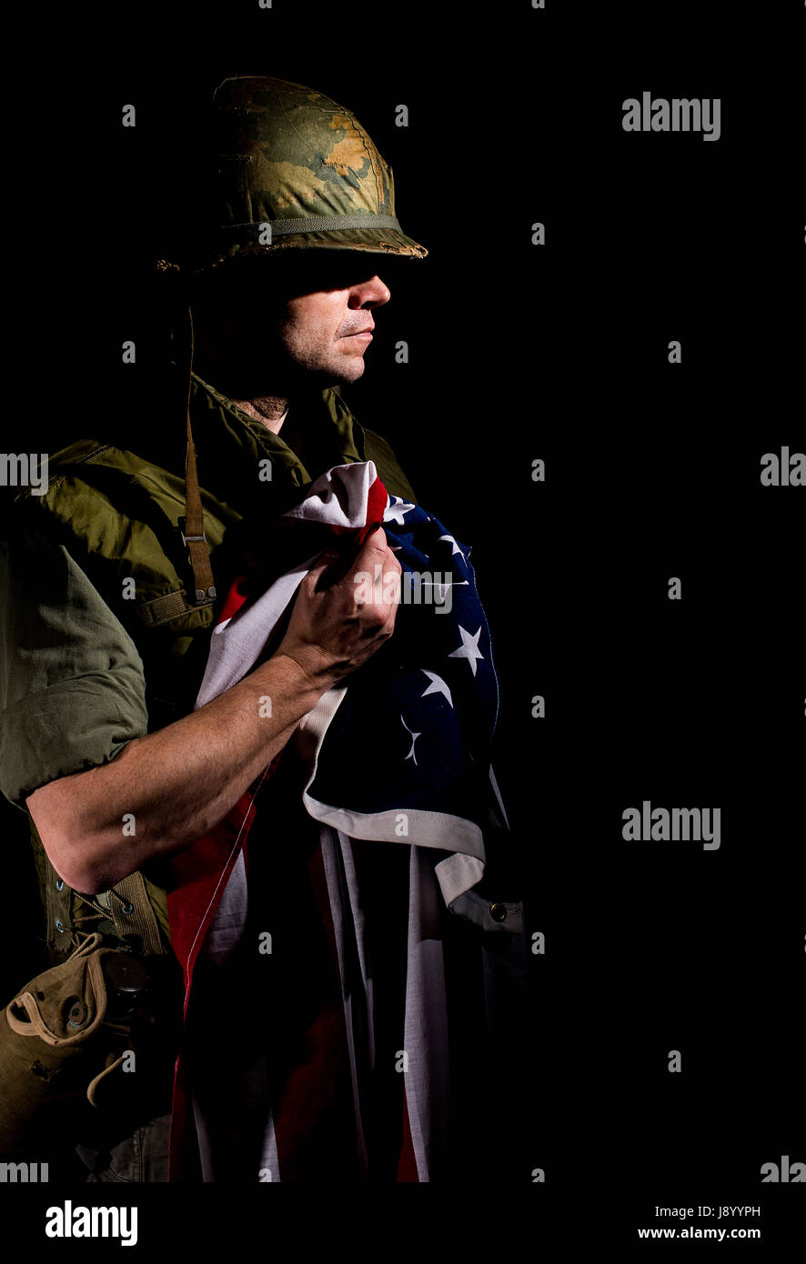 Patriotic US soldier from the Vietnam War period, holding the American flag tightly and standing against a dark background. Stock Photo