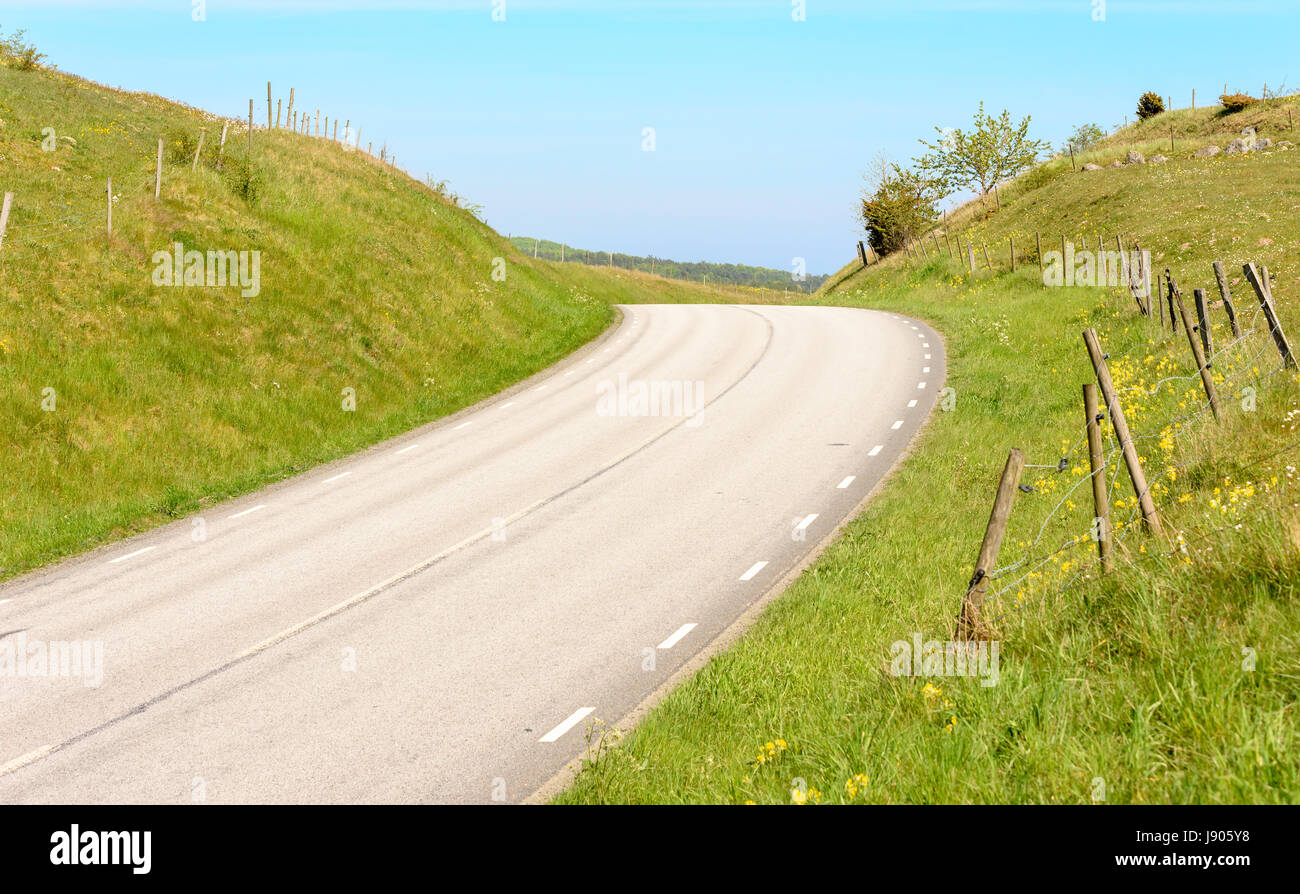 Narrow country road disappearing over the slope in the valley. Green pasture and fencing on either side of the road. Location Brosarp in Scania, Swede Stock Photo