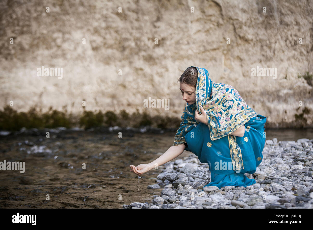 The girl in the Indian sari sits at the small river and drinks from ...