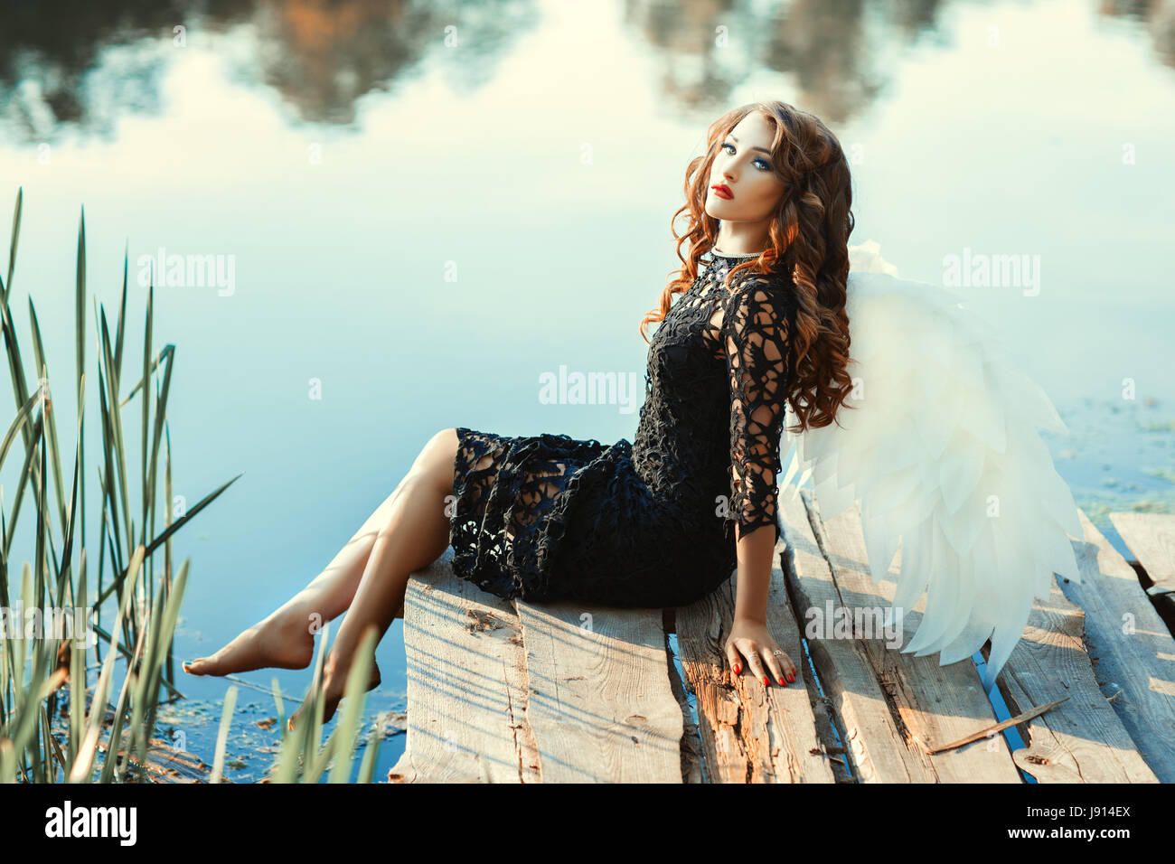 Girl with wings of an angel sitting near a river. She's in a black dress. See more photos of this series. Stock Photo