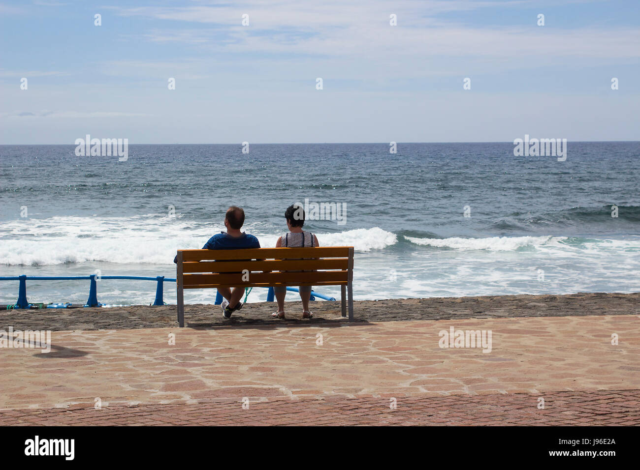 Holidaying couple sit and relax on a wooden bench as they look out into the Atlantic Ocean from the promenade at Playa Las Americas in Teneriffe in th Stock Photo