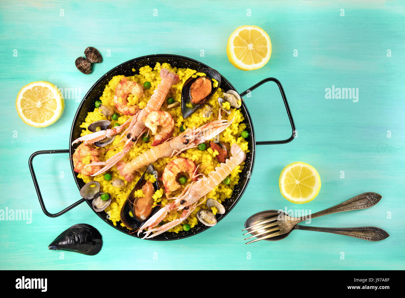 Photo of Spanish seafood paella in typical paellera, with slices of lemons, mussels and clams shells, fork and spoon, and a place for text, shot from  Stock Photo