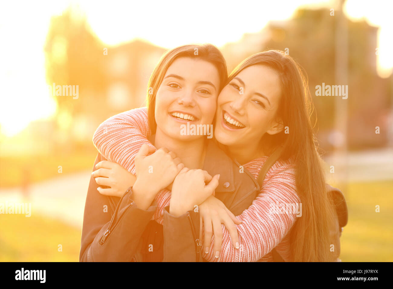 Front view portrait of two happy friends laughing and posing looking at you in the street at sunset with a warm light in the background Stock Photo